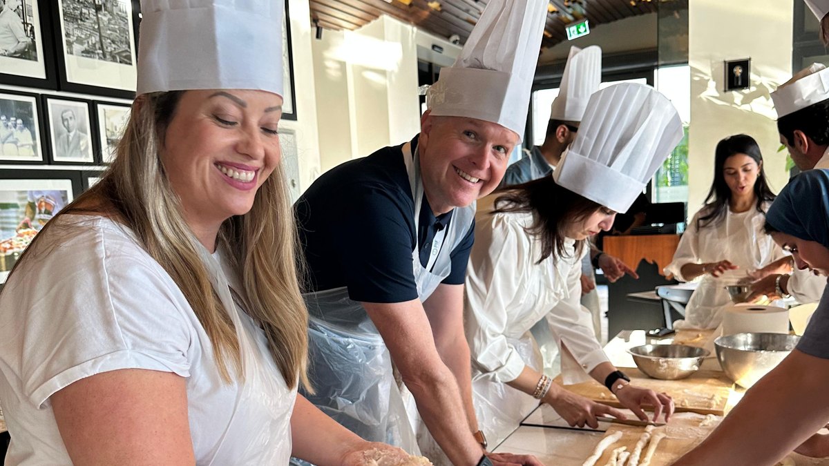 Two colleagues in chef hats laughing while they shape fresh pasta by hand