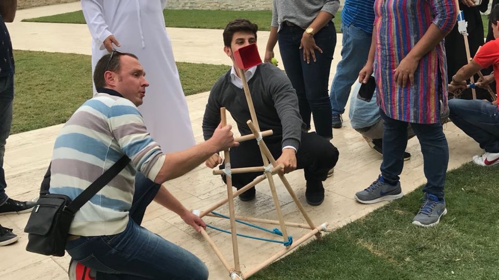 A participant kneeling on the grass to test a wooden catapult while the team watches