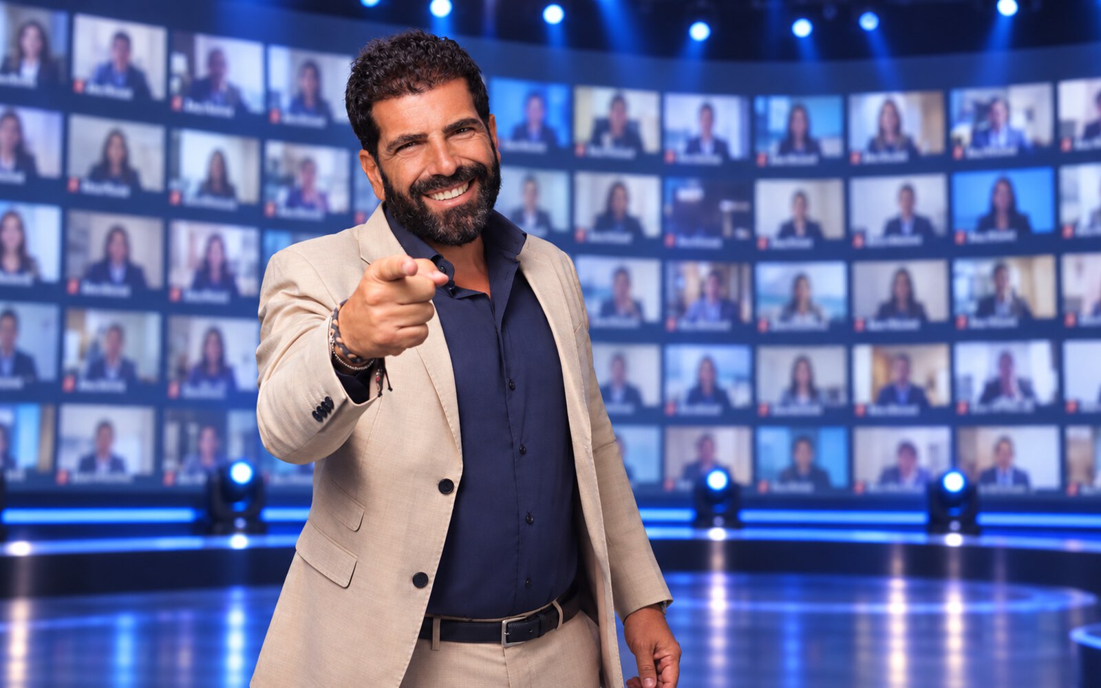 A host on stage in front of a wall of screens with the connected teams
