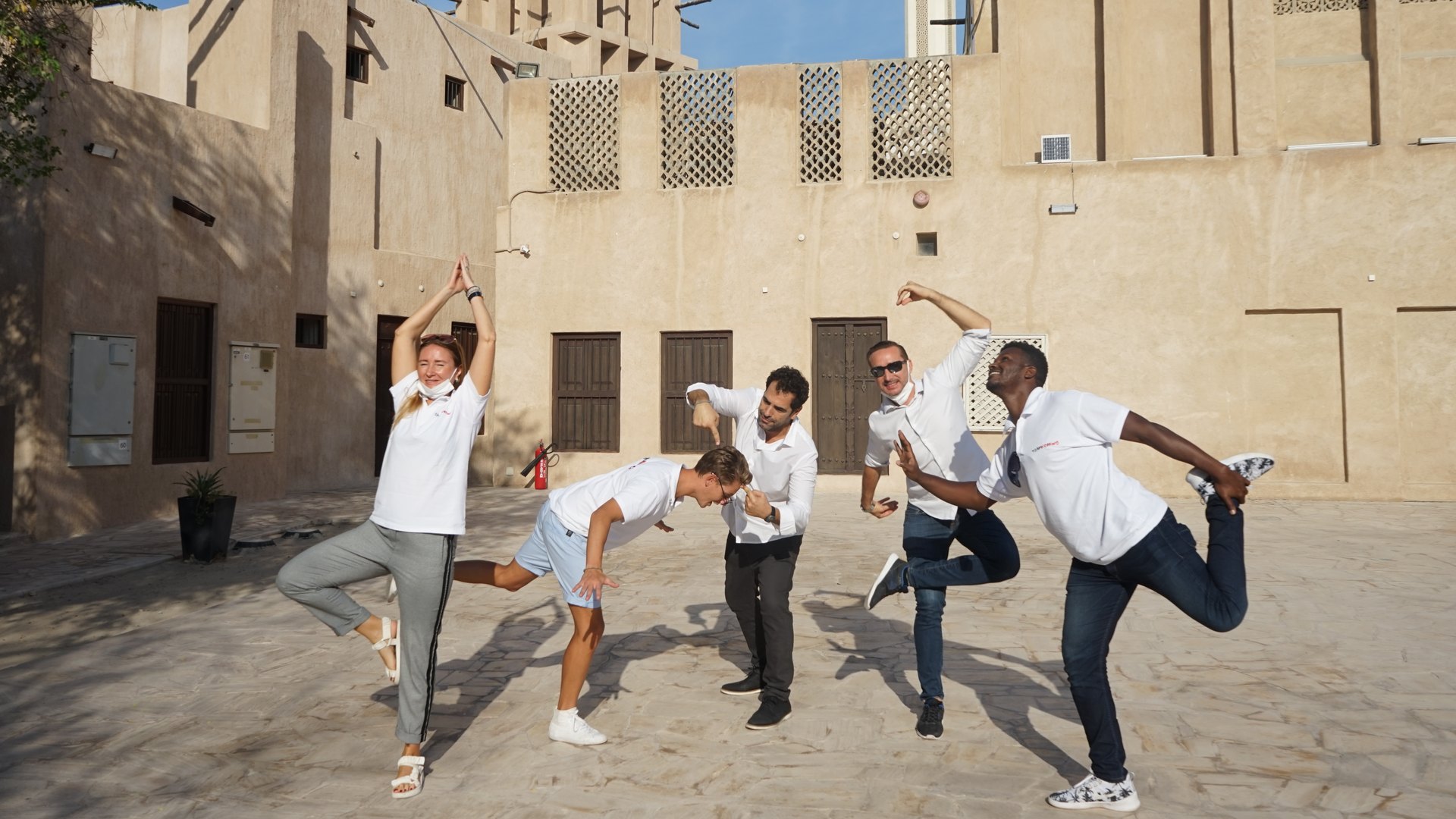 A team spread across a courtyard in the old quarter, each one in a different pose, a wind tower behind